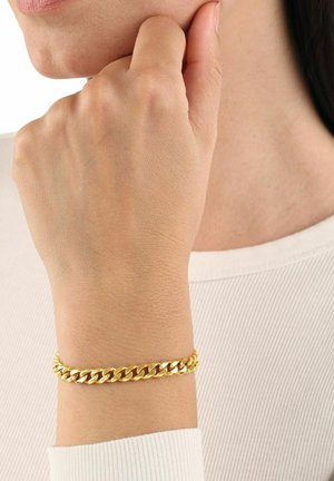 Bracciale a catena dorata indossato su un polso, con anelli intrecciati e una finitura lucida. Abbinato a un top bianco dalla texture.