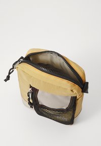 Gul stoffpose med en svart nettinglomme, sølvglidelås og oransje søm. Åpen topp som avdekker et lysegrått interiør. Kompakt design.