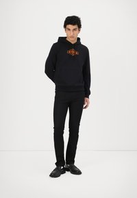 Svart hettegenser med oransje grafisk design, kombinert med svarte slim-fit bukser og svarte sko. Enkle, ensfargede farger gjennom hele antrekket.