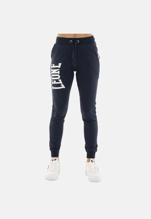 Pantalon de survêtement bleu marine avec texte blanc « LEONE » sur la cuisse gauche, taille à cordon, chevilles resserrées, porté avec des baskets blanches.