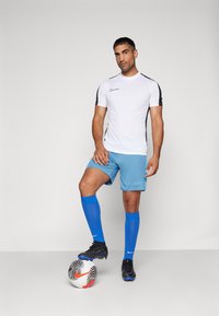 Balta Nike sportinė marškinėliai su juodais akcentais, mėlynos šortai, mėlynos kojinės ir juodi futbolo batai. Futbolo kamuolys yra atsidūręs prie kojų.