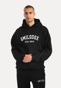 Zwarte hoodie met witte tekst "SMILODOX ATHL. WEAR". Gemaakt van zacht materiaal, met een kangoeroezak en geribbelde boorden.