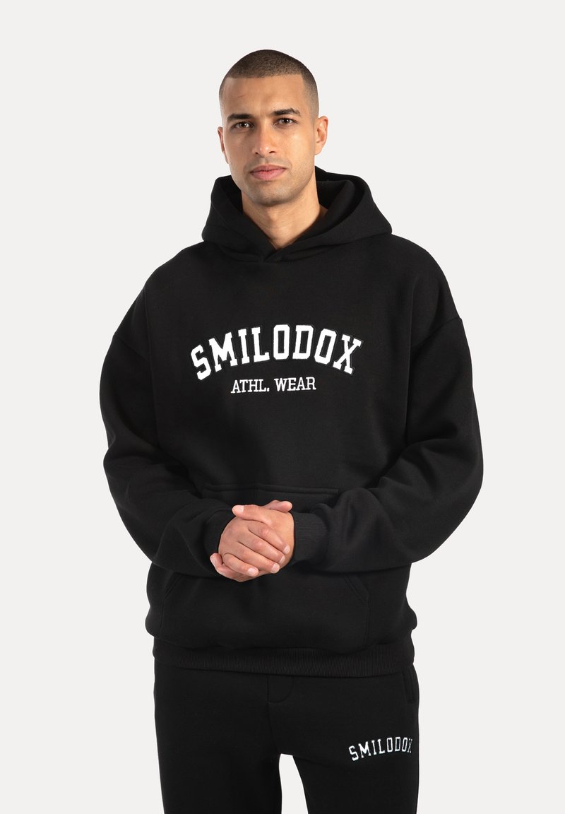 Zwarte hoodie met witte tekst "SMILODOX ATHL. WEAR". Gemaakt van zacht materiaal, met een kangoeroezak en geribbelde boorden.