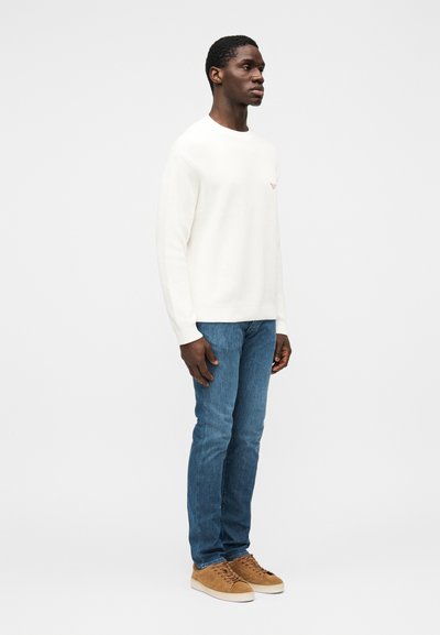 Witte gebreide trui met een ronde hals, gecombineerd met blauwe spijkerbroek. Het model draagt beige suède sneakers en staat tegen een grijze achtergrond.