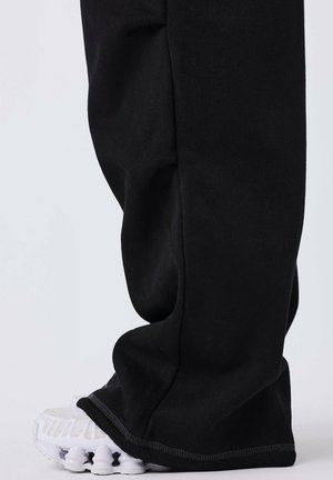 Pantalons de survêtement noirs à coupe décontractée, se terminant juste au-dessus d'une sneaker blanche avec une semelle texturée et un design distinctif.