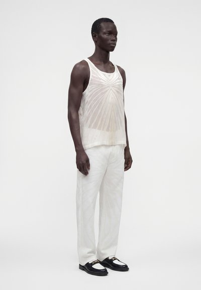Homme portant un débardeur blanc translucide à motifs, un pantalon blanc ample et des mocassins noirs et blancs, debout devant un fond uni.