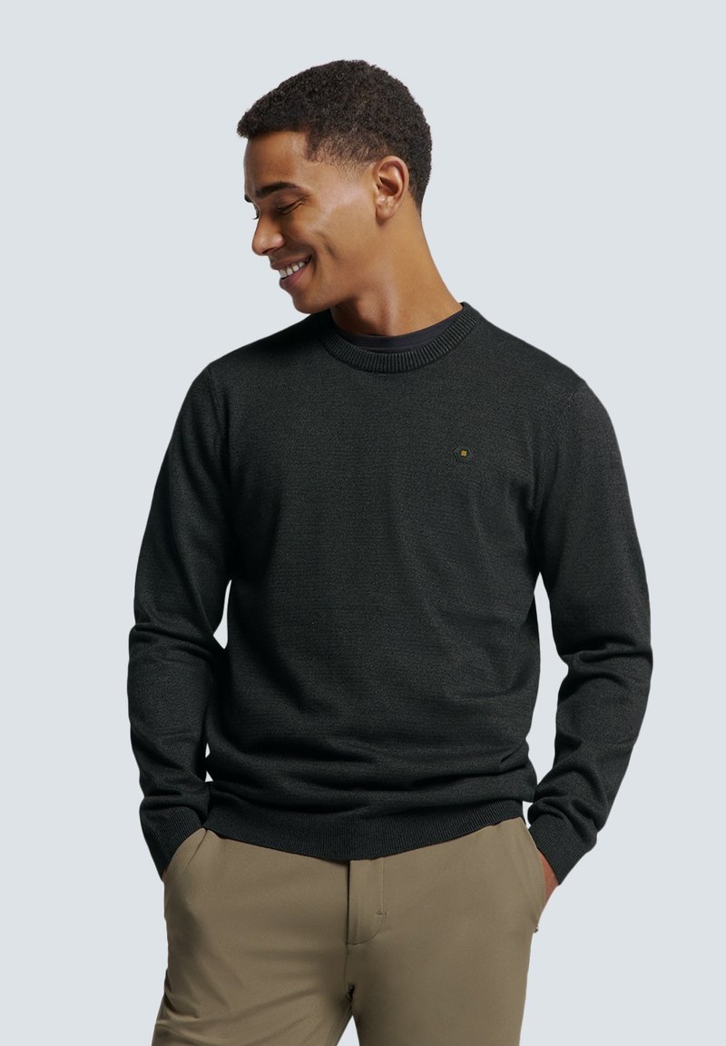 Pull en maille gris foncé avec un col ras du cou, des poignets côtelés et un petit logo sur la poitrine, associé à un pantalon de couleur claire.