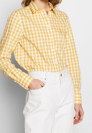 Koszula w żółto-białą kratę na guziki, z długimi rękawami i kołnierzykiem. W połączeniu z białymi spodniami. Tkanina wydaje się lekka i gładka w dotyku.