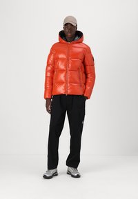 Orange puffer jacket with a glossy finish, zip closure, and hood. Paired with black cargo pants and beige cap, featuring gray sneakers.