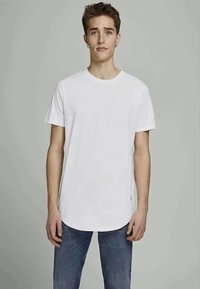 Jovem de pé, virado para a frente, vestindo uma camiseta branca simples e jeans azuis, em frente a um fundo cinza neutro.