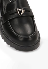 Mocasín de cuero negro con un detalle metálico en forma de corazón brillante en una correa. Cuenta con un upper texturizado y una suela de goma gruesa.