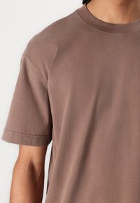 Brown cotton t-shirt with a round neck, short sleeves, and a relaxed fit. Smooth texture with visible stitching details on sleeves.