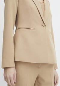 Beige blazer met notch revers, enkele knoopsluiting en zijzakken. Glad textuur met getailleerde pasvorm. Bijpassende broek zichtbaar.