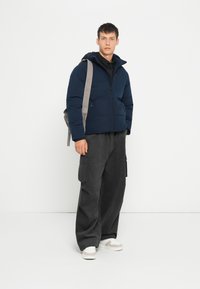 Marineblå pufferjakke med hætte, lynlås og sidelommer. Sammen med grå cargobukser og beige sneakers. Minimalistisk design.