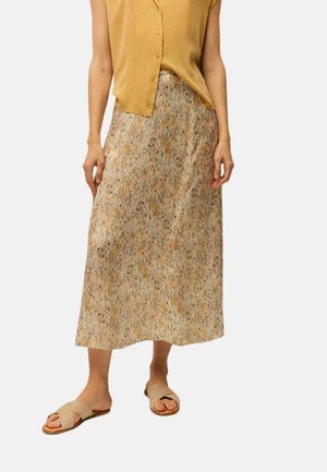 Mujer con un top sin mangas abotonado de color mostaza, falda midi beige con estampado floral y sandalias planas beige tejidas, de pie sobre un fondo blanco.