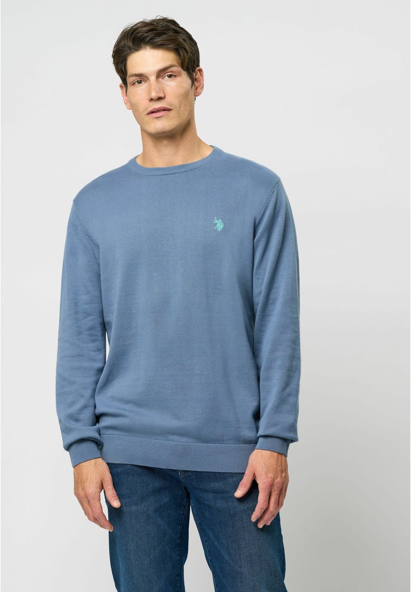 Blå strikket sweater med rund hals og ribbede manchetter. Har et lille turkist logo på brystet, parret med jeans.
