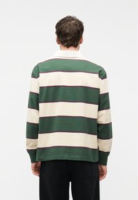 Άνδρας που φαίνεται από πίσω, φοράει μακρυμάνικο πουκάμισο ράγκμπι με φαρδιές οριζόντιες πράσινες, κρεμ και λεπτές μωβ ρίγες, και μαύρο παντελόνι.