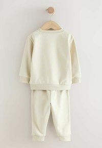 Ensemble de survêtement en coton couleur crème avec un haut à manches longues et un pantalon. Présente des poignets et un ourlet côtelés, une texture lisse et un design simple.