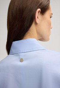 Camicia azzurro chiaro con una texture liscia, caratterizzata da un dettaglio con bottone logo argentato sul colletto posteriore. Vista ravvicinata di lato.