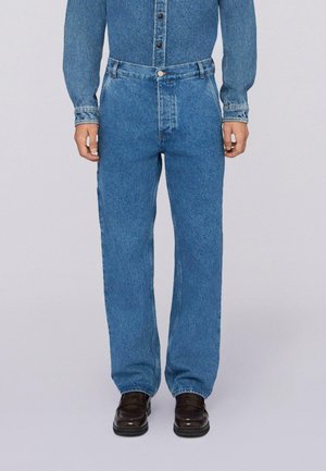 Hombre vestido con jeans de mezclilla azul y una camisa de mezclilla azul a juego, combinados con zapatos de cuero marrón oscuro, de pie frente a un fondo neutral.