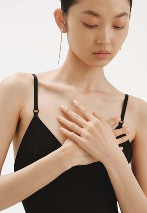 Top negro sin mangas con tirantes finos, que presenta una textura suave. Las manos del modelo están cruzadas sobre el pecho, mostrando dedos delicados y pendientes.