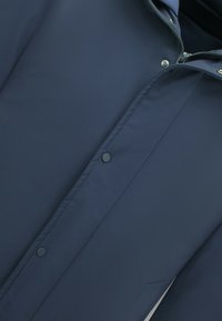 Marineblauwe jas met een gladde, waterdichte stof, een rits aan de voorkant en drukknoopsluiting. Voorzien van een capuchon en zijzakken voor functionaliteit.