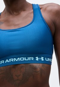 Persoană musculoasă purtând un top sportiv albastru Under Armour, arătând trunchiul superior și un tatuaj cu leu pe partea laterală a pieptului.