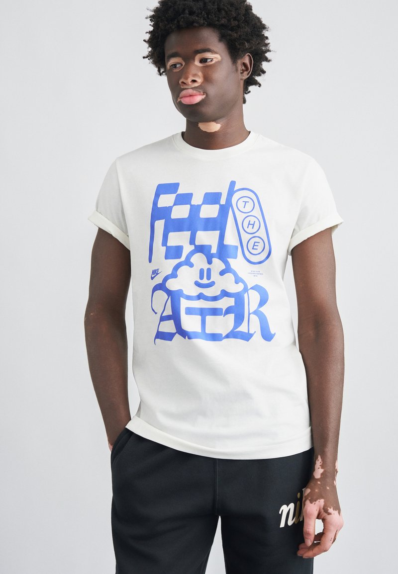 Jeune homme atteint de vitiligo portant un t-shirt blanc avec un imprimé graphique bleu et un pantalon noir, debout devant un fond clair uni.