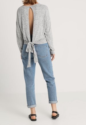 Femme portant un haut à manches longues gris clair avec un dos ouvert noué à la taille, un jean bleu roulé et des sandales noires, debout devant un fond blanc.