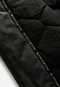 Schwarze gesteppte Pufferjacke mit glatter Textur, geometrischem Muster und einem markierten Reißverschluss mit "ALLSUNITS" in Weiß bedruckt.