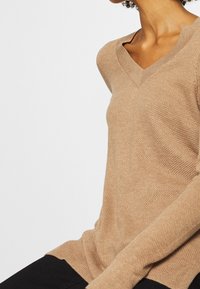 Personne portant un pull beige texturé à col en V et un pantalon noir, assise devant un fond gris clair uni.