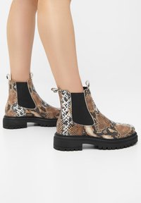 Bottes chevilles à motif peau de serpent avec panneaux latéraux élastiques noirs, accents argentés et semelle épaisse en caoutchouc noir. Détail de surface texturée visible.