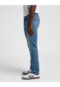 Jeans de denim azul en un diseño de corte recto, con cinco bolsillos y detalles de costura visibles. Combinados con zapatillas blancas con rayas negras.