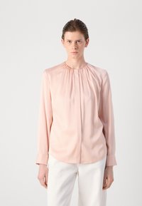 Blusa rosa chiaro a maniche lunghe con scollo rotondo arricciato e delicate pieghe davanti, realizzata in tessuto morbido. Pantaloni bianchi.