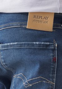 Jeans de denim azul con una gran etiqueta de cuero marrón claro con "REPLAY HYPERFLEX" estampado. Los detalles incluyen costuras y un pequeño acento rojo.