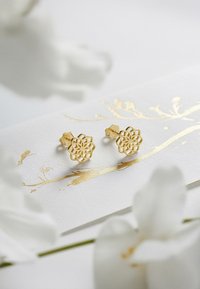 Goldene florale Ohrstecker mit filigranem Ausschnittdesign. Auf einer weißen Karte montiert, die goldene Blattelemente und einen sanften floralen Hintergrund aufweist.