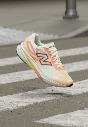 Biały i pomarańczowy but do biegania New Balance w powietrzu nad szarym przejściem dla pieszych na asfaltowej drodze.