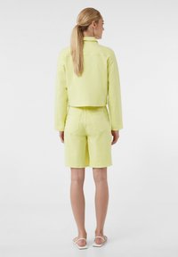 Jasnożółta dżinsowa kurtka o skróconym kroju oraz dopasowane szorty, z standardowymi kieszeniami i schludnym wykończeniem. Całość dopełniają płaskie sandały.