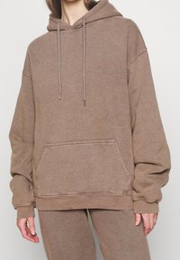 Brązowa, oversize'owa bluza z kapturem wykonana z miękkiego materiału, posiadająca przednią kieszeń typu kangur, kaptur ze ściągaczem oraz subtelne haftowane logo na piersi.