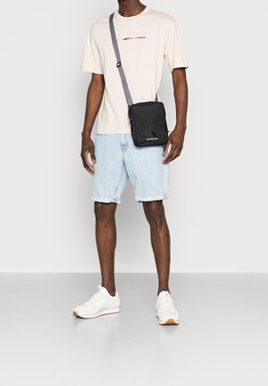 Mand iført hvide sneakers, lyseblå denimshorts, beige t-shirt med farvet logo og sort skuldertaske med grå rem.