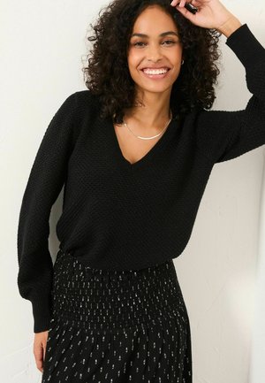 Schwarzer, gestrickter Pullover mit V-Ausschnitt und langen Ärmeln, kombiniert mit einem schwarzen Rock mit gerafftem Taillenbund und weißen Mustern.