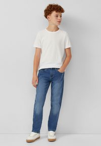 Bela bombažna majica, modri kavbojasti hlače z rahlim bledenjem in bele superge s svetlo bež podplati, stoječa na nevtralni ozadju.