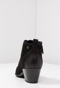 Botas tobilleras negras de ante con un pequeño tacón, que cuentan con una lengüeta trasera y un acento metálico cerca de la parte superior. Textura suave y líneas limpias.