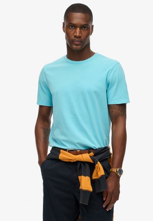 T-shirt en coton bleu clair avec un col rond, manches courtes, associé à un pantalon bleu marine et un pull rayé jaune et gris noué autour de la taille.