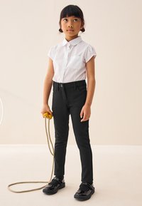 Camicia bianca a collo, pantaloni neri aderenti, scarpe nere lucide e una corda per saltare gialla. Tessuto liscio, vestibilità slim e design pratico.