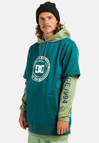 Turkusowy, oversize'owy hoodie z zielonymi warstwowymi rękawami, przednią kieszenią i okrągłym białym logo. Posiada sznurki oraz wyraźny tekst na rękawach.