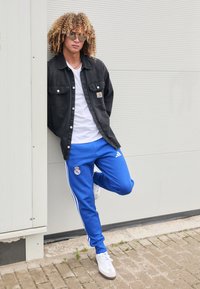 Black denim jacket over a white t-shirt, blue athletic pants with white stripes and logo, gray and white sneakers, standing against a wall.