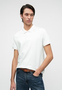 Cămașă polo albă cu guler clasic, mâneci scurte și deschidere cu nasturi; asortată cu blugi albaștri și o curea maro.