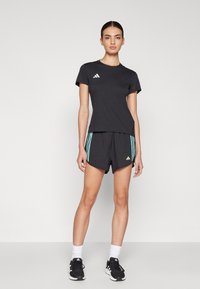 Camiseta deportiva negra de mangas cortas con logo blanco, combinada con pantalones cortos negros con rayas turquesa, llevada con zapatillas negras y calcetines blancos.