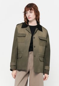 Casaco verde-azeitona com colarinho de veludo cotelê preto, dois bolsos no peito e corte reto. Fecho com botões e material texturizado.